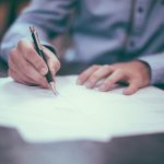 man in a button-up shirt writes on papers with a black pen